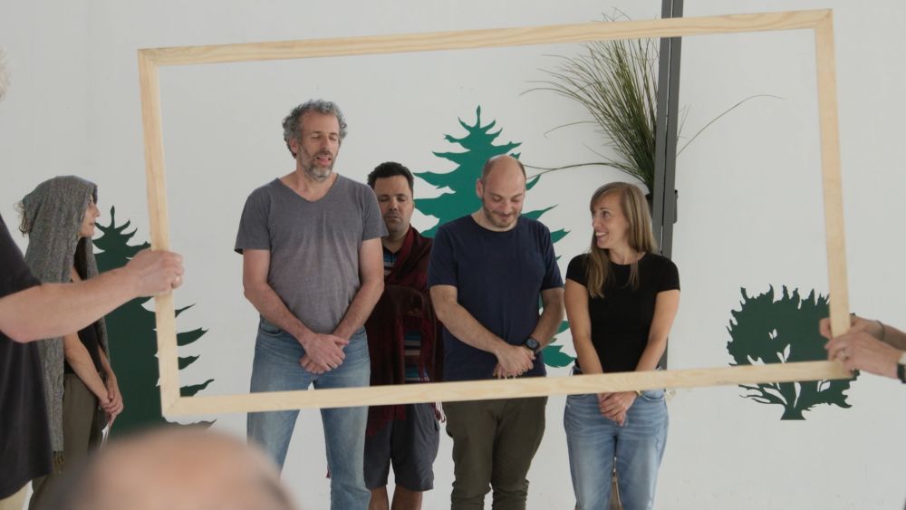 Záber z filmu Return to Epipo: štyri dospelé osoby stoja pred stenou, ďalší ľudia pred nimi držia drevený rám